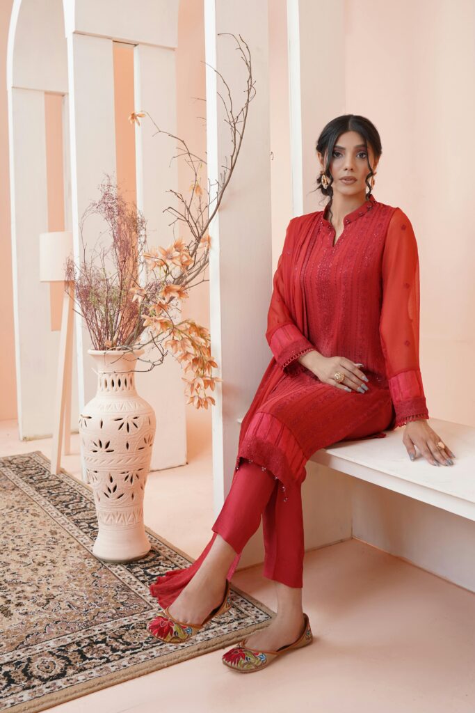 A woman in traditional red clothing sitting elegantly indoors, showcasing Pakistani fashion.