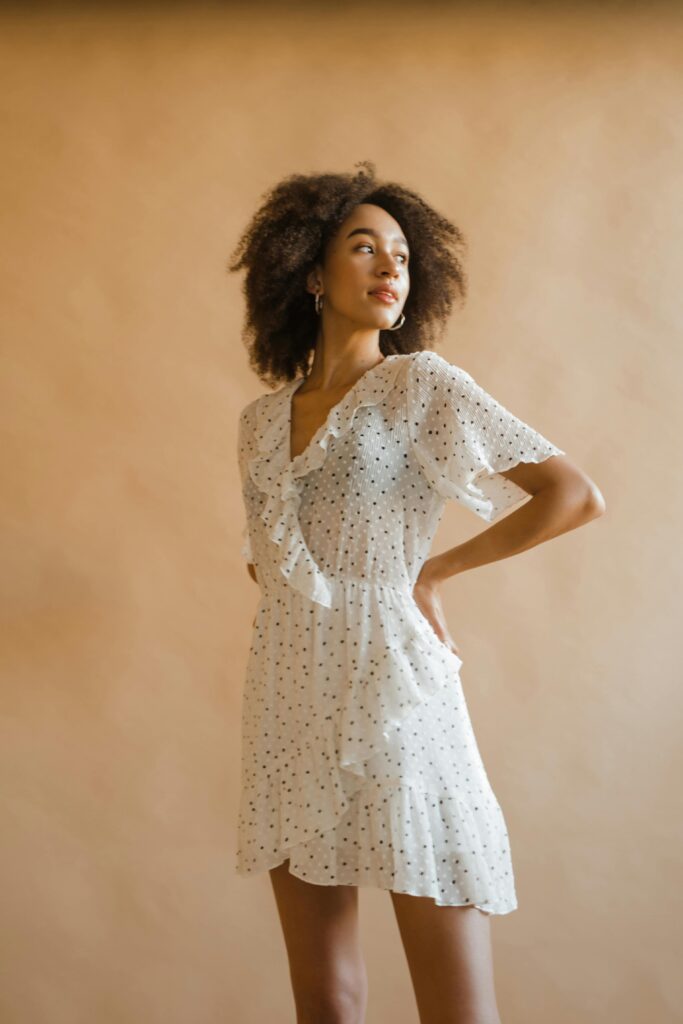 Fashionable woman in a white dress posing confidently against a warm, neutral background.